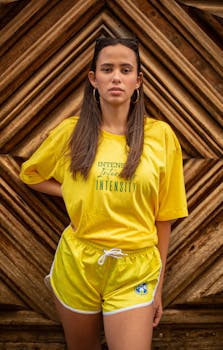 A woman in a yellow shirt and shorts standing outdoors against a wooden backdrop.