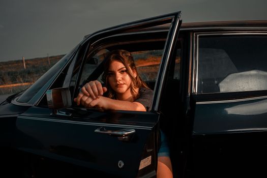 A woman leans out of a classic car door, embracing a serene sunset moment outdoors.