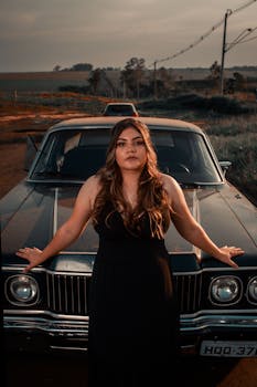 A fashionable woman in a black dress poses confidently against a vintage car during sunset on a country road.