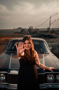 A smiling woman in a black dress posing with a vintage car on a countryside road.