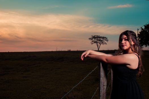 A woman with long hair poses by a fence in the countryside during sunset, creating a serene scene.