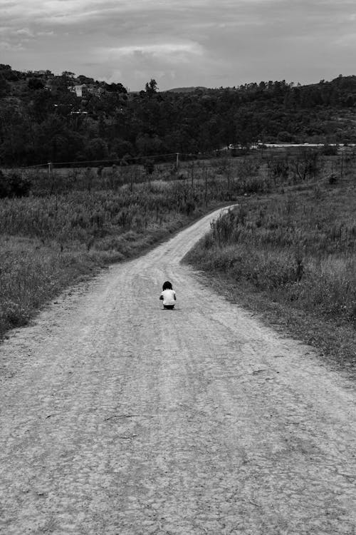 Grayscale Photo of a Child Sitting in the Middle of a Dirt Road