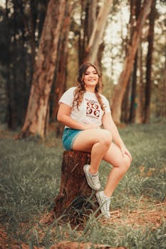 A woman sitting casually on a tree stump in a forest, smiling and enjoying the serene outdoor environment.