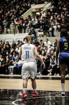 Two female basketball players competing during a lively match with spectators in the background.