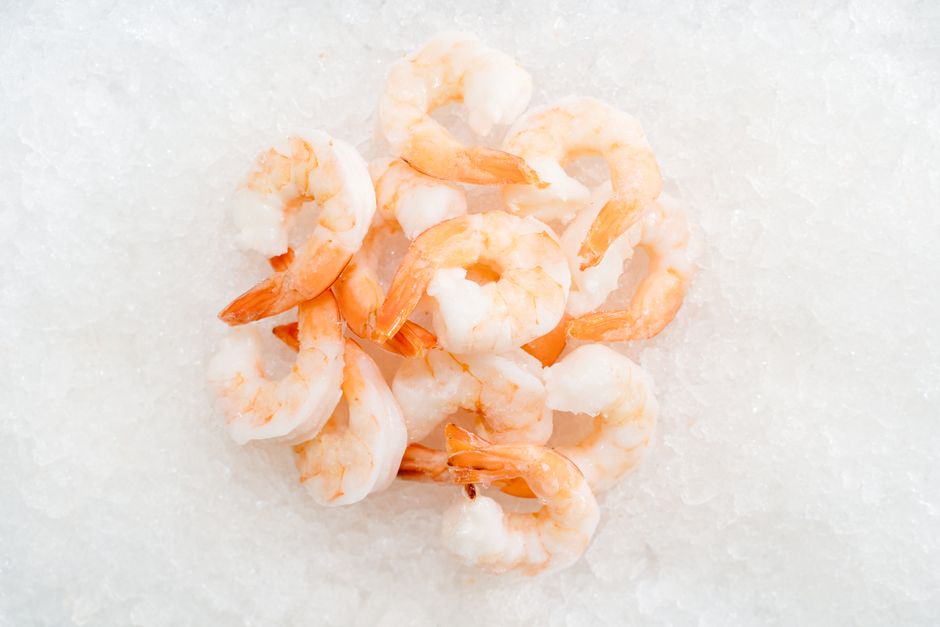 A pile of raw, peeled, tail-on frozen shrimp thawing on a bed of ice next to the shrink-wrapped bag showing a 21/25 count label.