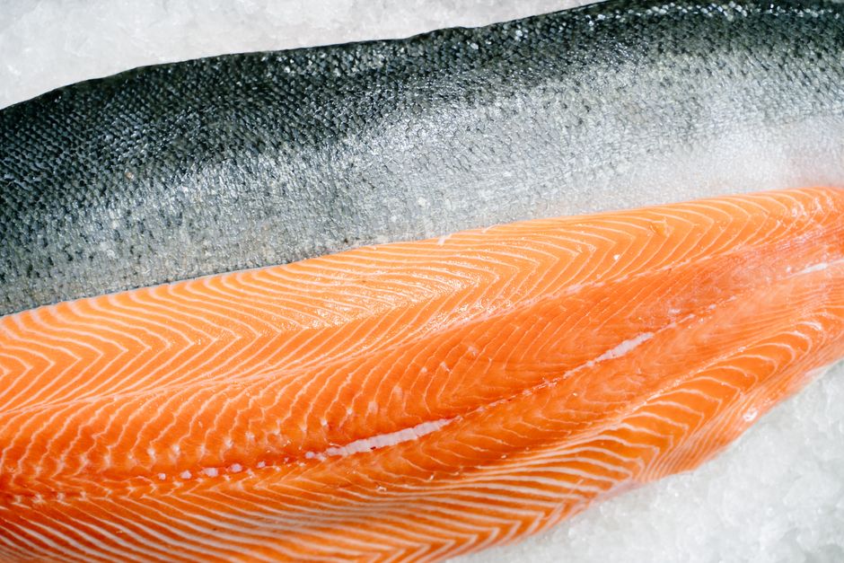 Two salmon fillets on a cutting board, one deep red-orange and leaner labeled wild-caught, the other paler orange with visible white fat marbling labeled farmed, next to a wedge of lemon.