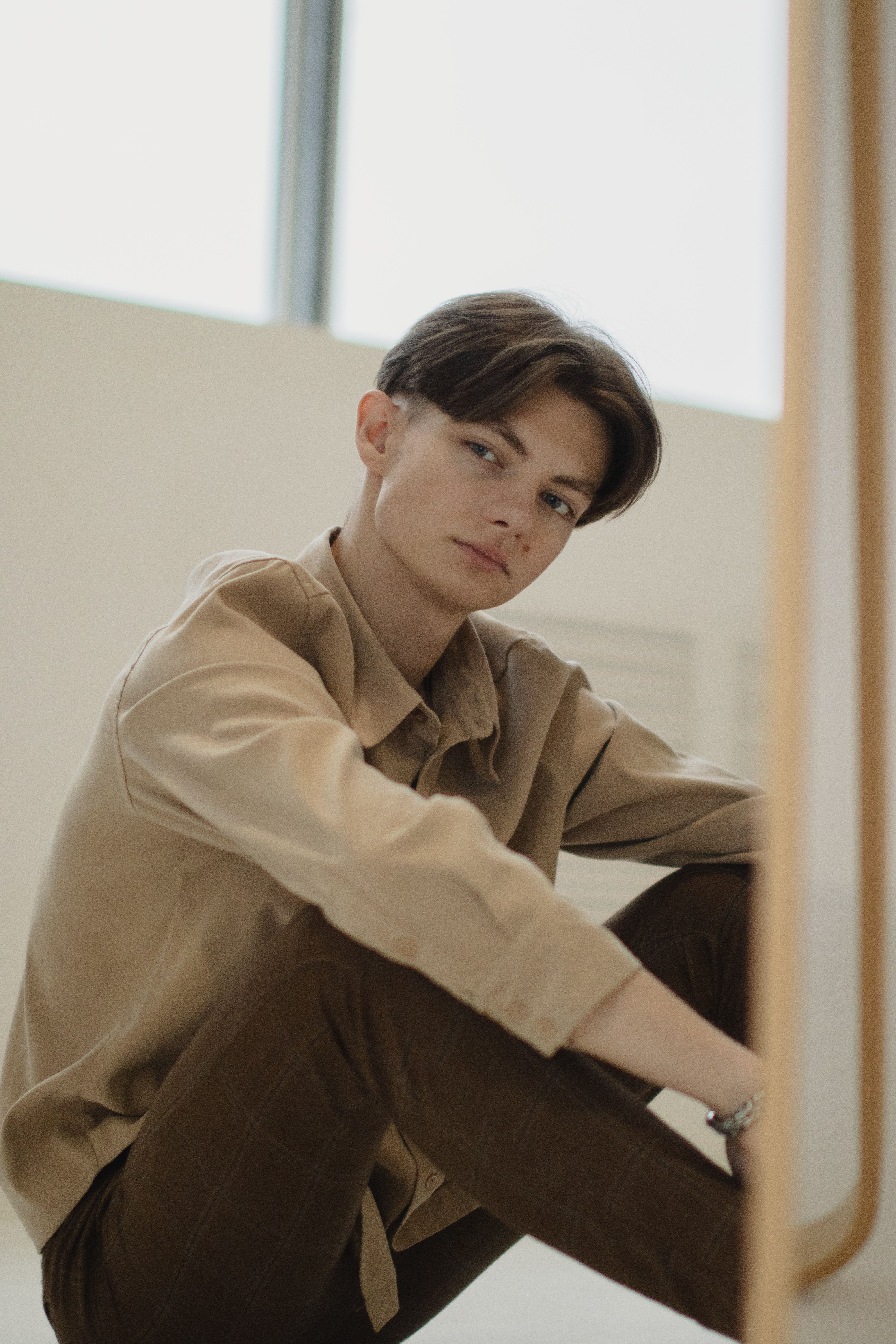 A contemplative young man in brown clothing sits near a mirror indoors.