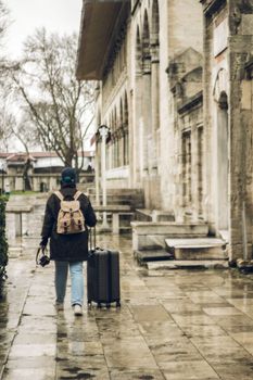 Person with backpack and luggage walking on a rainy day near a historic building.