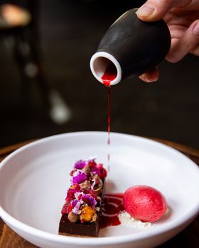 Close-up of a gourmet dessert with berry sauce being poured, featuring vibrant garnishes.
