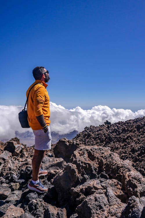 A Man in Orange Jacket Standing on Big Rocks Near Cloudy Sky