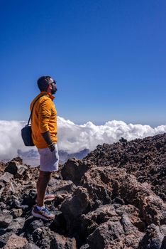 Man in orange jacket stands on rocky mountain peak under blue sky and clouds.