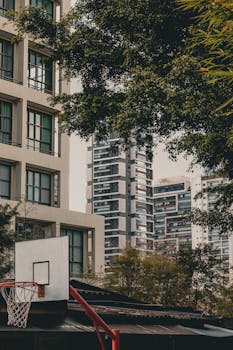 Basketball hoop in an urban park surrounded by high-rise buildings and lush greenery, capturing street sports vibe.