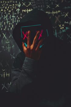 A person in a black hoodie with neon mask sits in a dark, moody setting surrounded by abstract symbols.