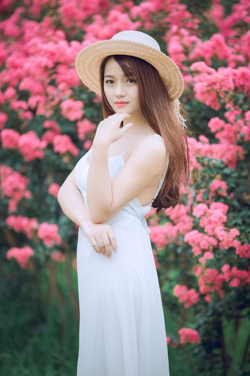 분홍색 꽃잎 꽃 앞에서 흰 드레스와 태양 모자를 입은 여성의 피사계 심도