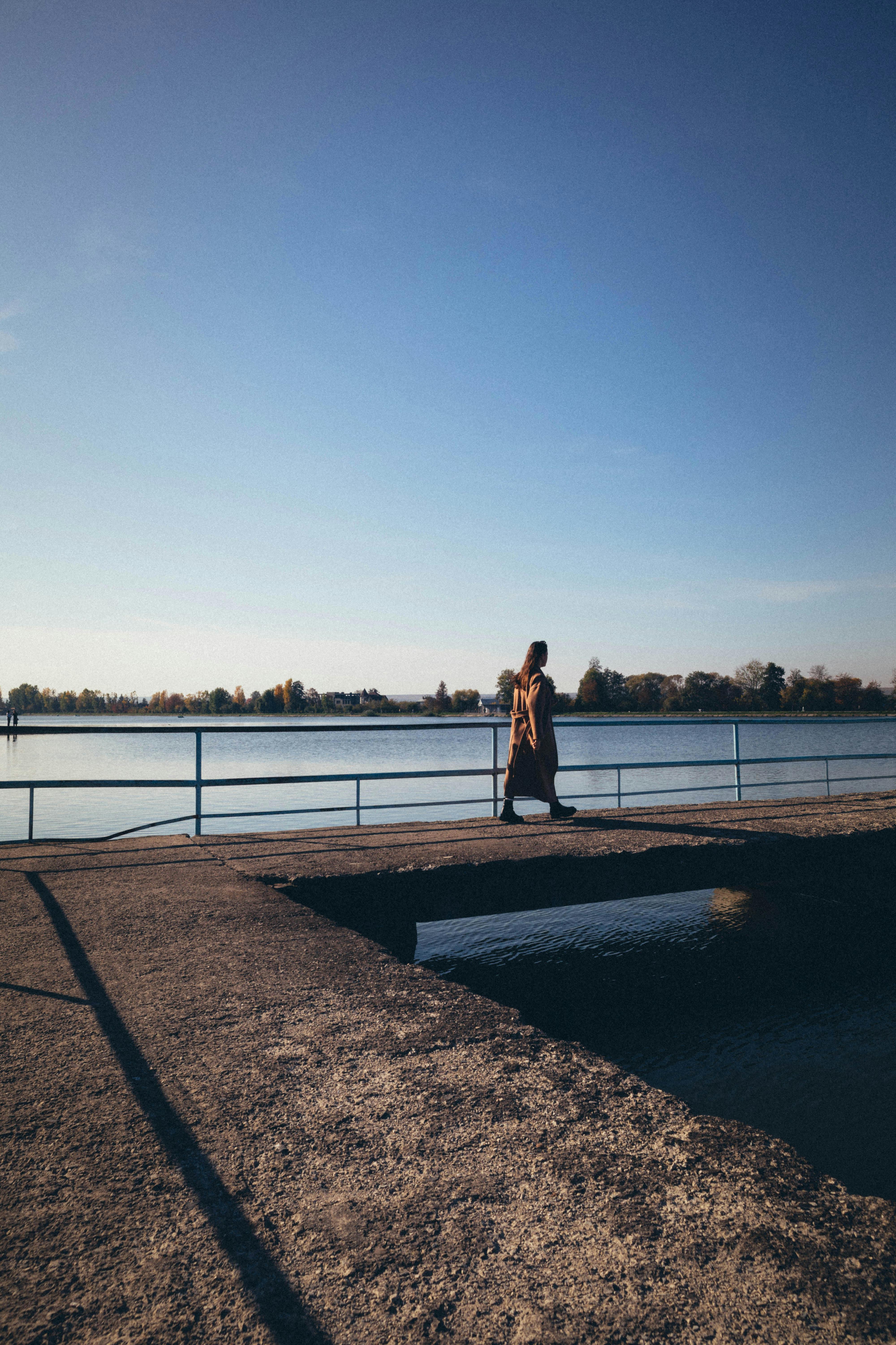 Free A woman walks along a dock under a clear blue sky near a calm lake, capturing a serene outdoor moment. Stock Photo
