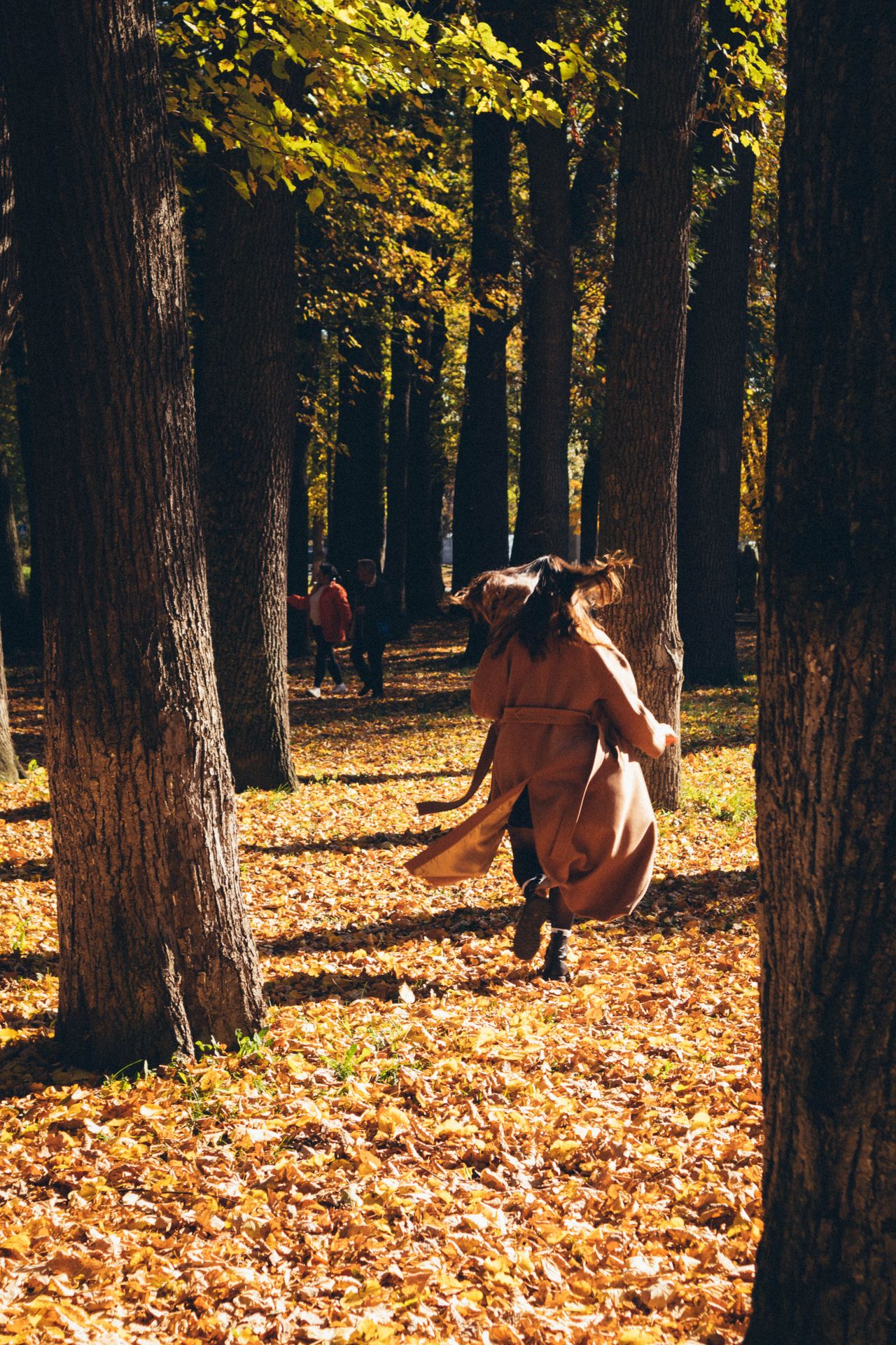 Woman in a tan trench coat walking through an autumn park with falling leaves