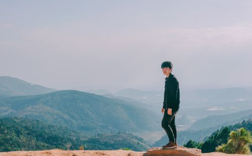 Free A young man stands on a mountain ridge enjoying a scenic landscape view under a clear sky. Stock Photo