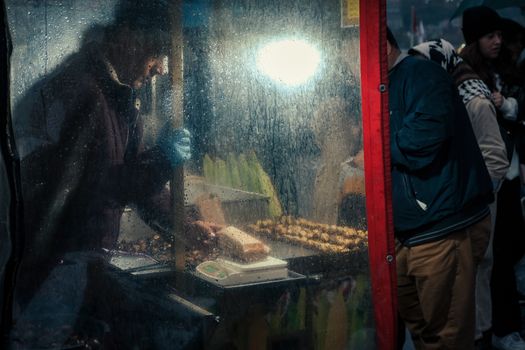A street food vendor prepares food at night behind a rainy window, serving people outdoors.