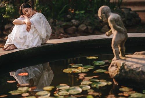 A Woman in White Dress Sitting Beside the Pond