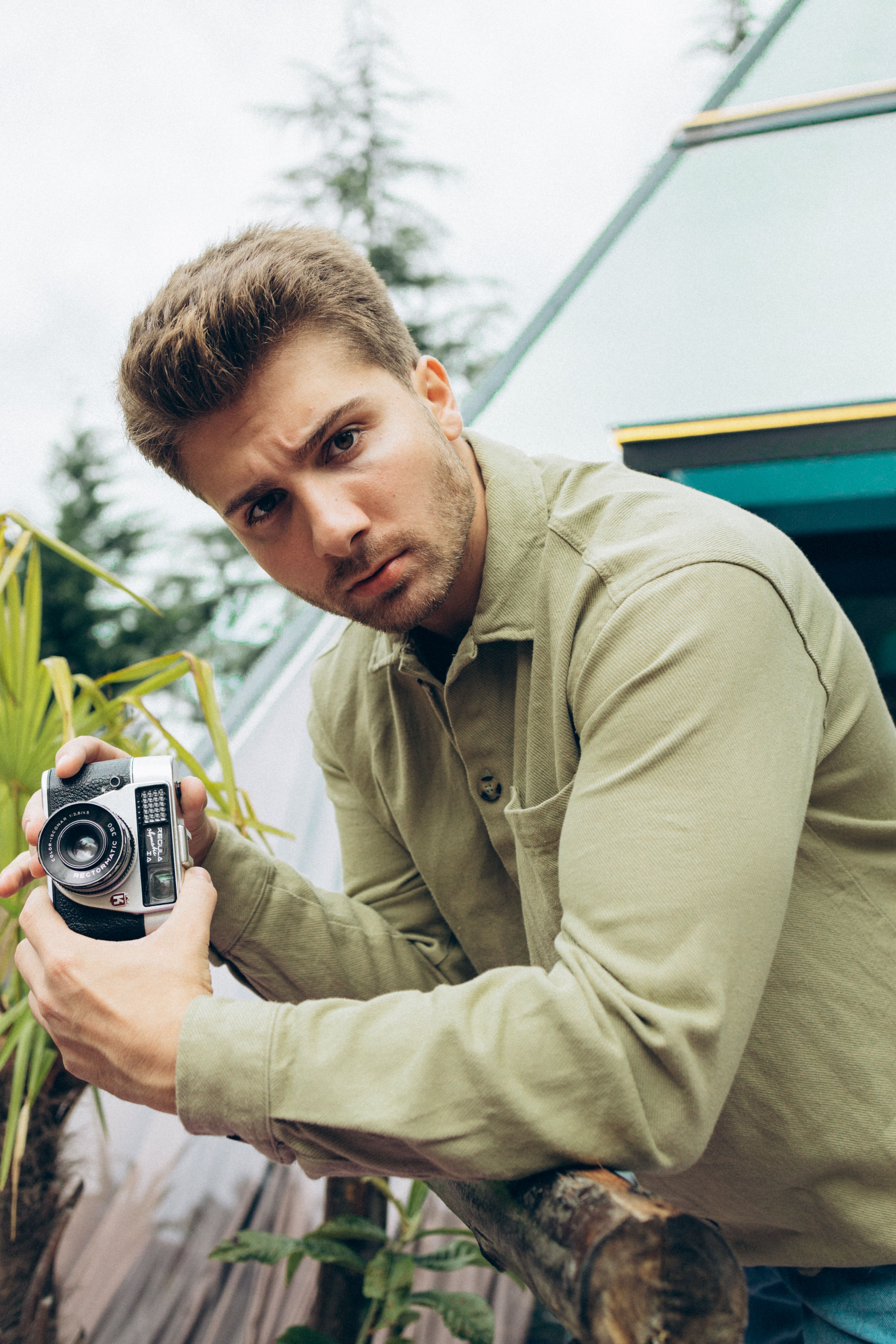 Young man holding a camera outdoors in Sapanca, Turkey, with greenery and modern architecture.