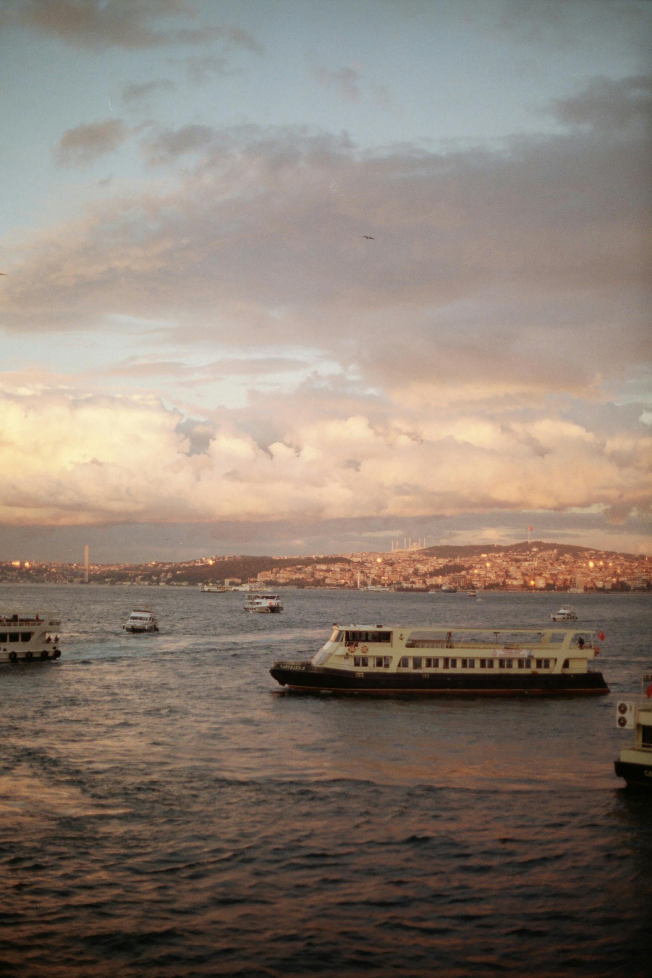 Passenger ships sail along a tranquil urban waterway at sunset, under a picturesque sky.