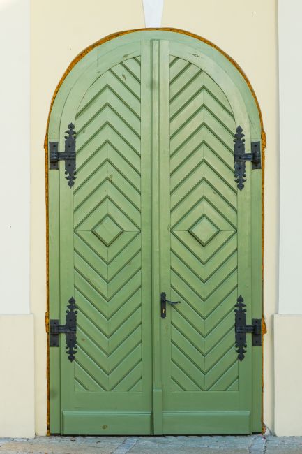 A tall, green, wooden door with intricate vintage-style black hinges on a pale wall.