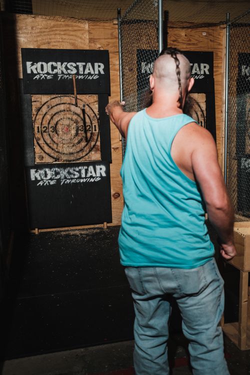 Free A man in a blue tank top engages in axe throwing at an indoor range with wooden targets. Stock Photo