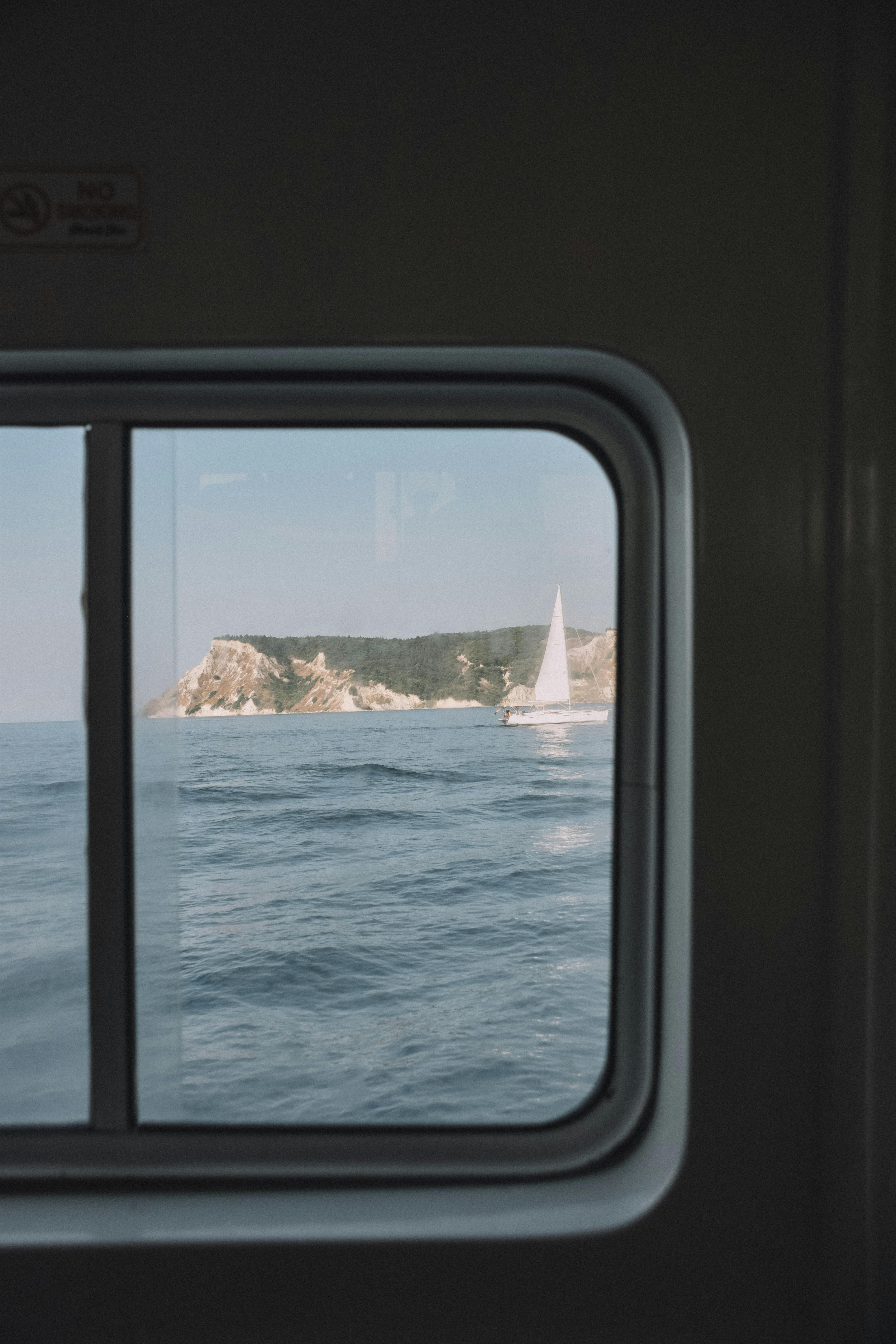 A serene window view of a sailboat and coastline from a ferry in Corfu, Greece.