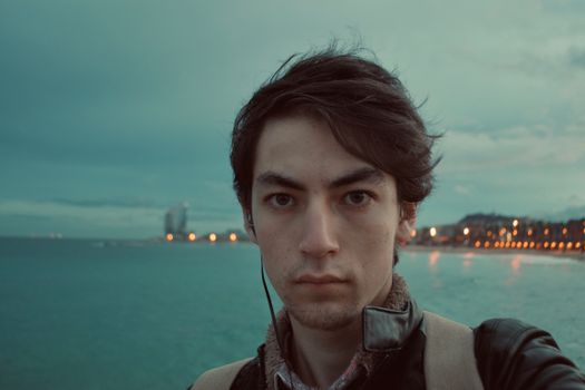 Portrait of a young man at the waterfront with city lights in the background during twilight.