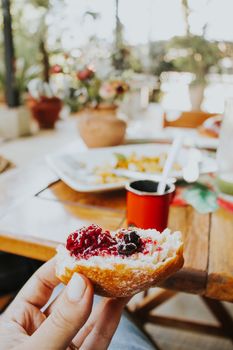 Close-up of a hand holding a brioche with jam, set on an outdoor breakfast table.