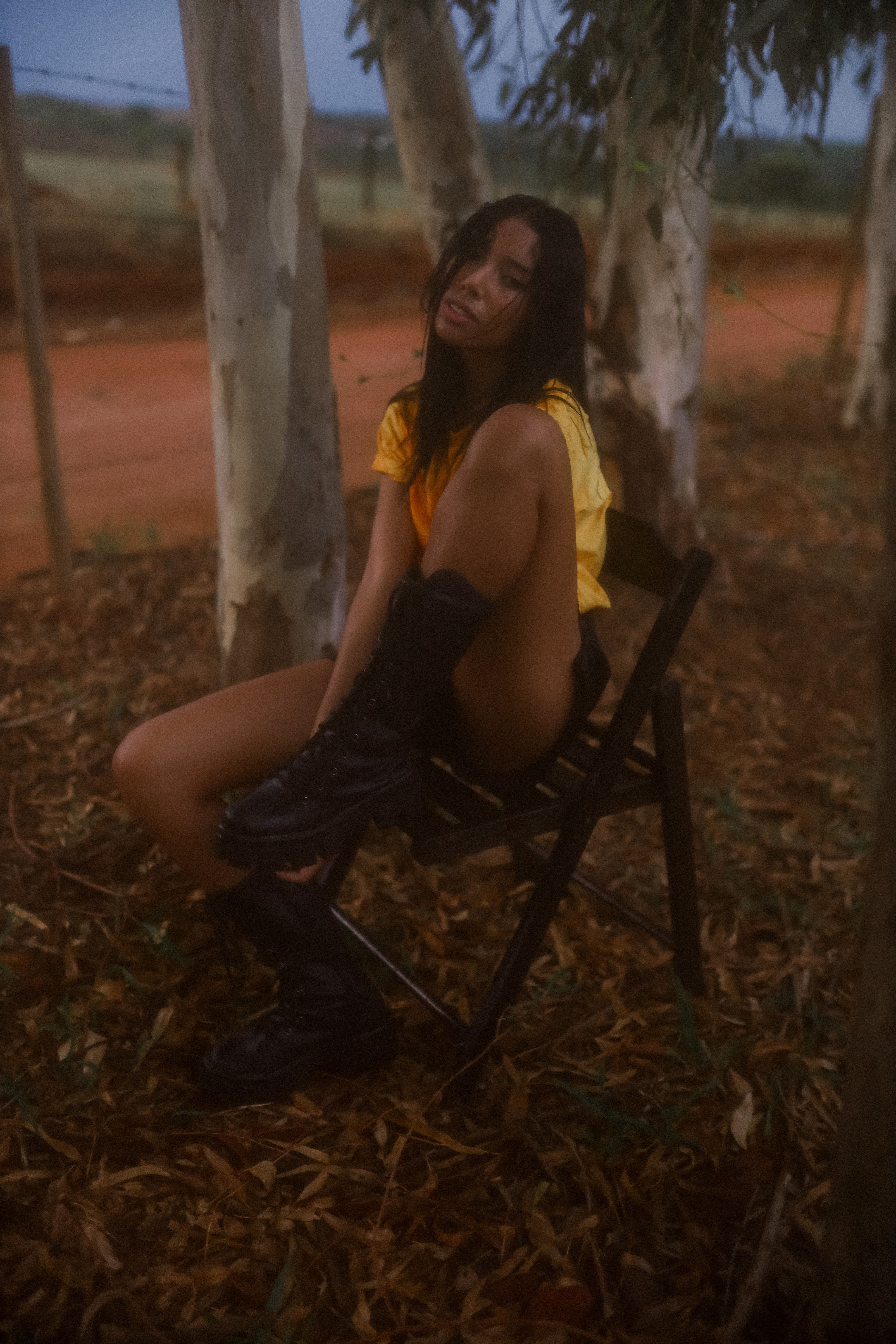 A woman in a yellow top and black boots sits on a chair amidst fallen leaves outdoors.