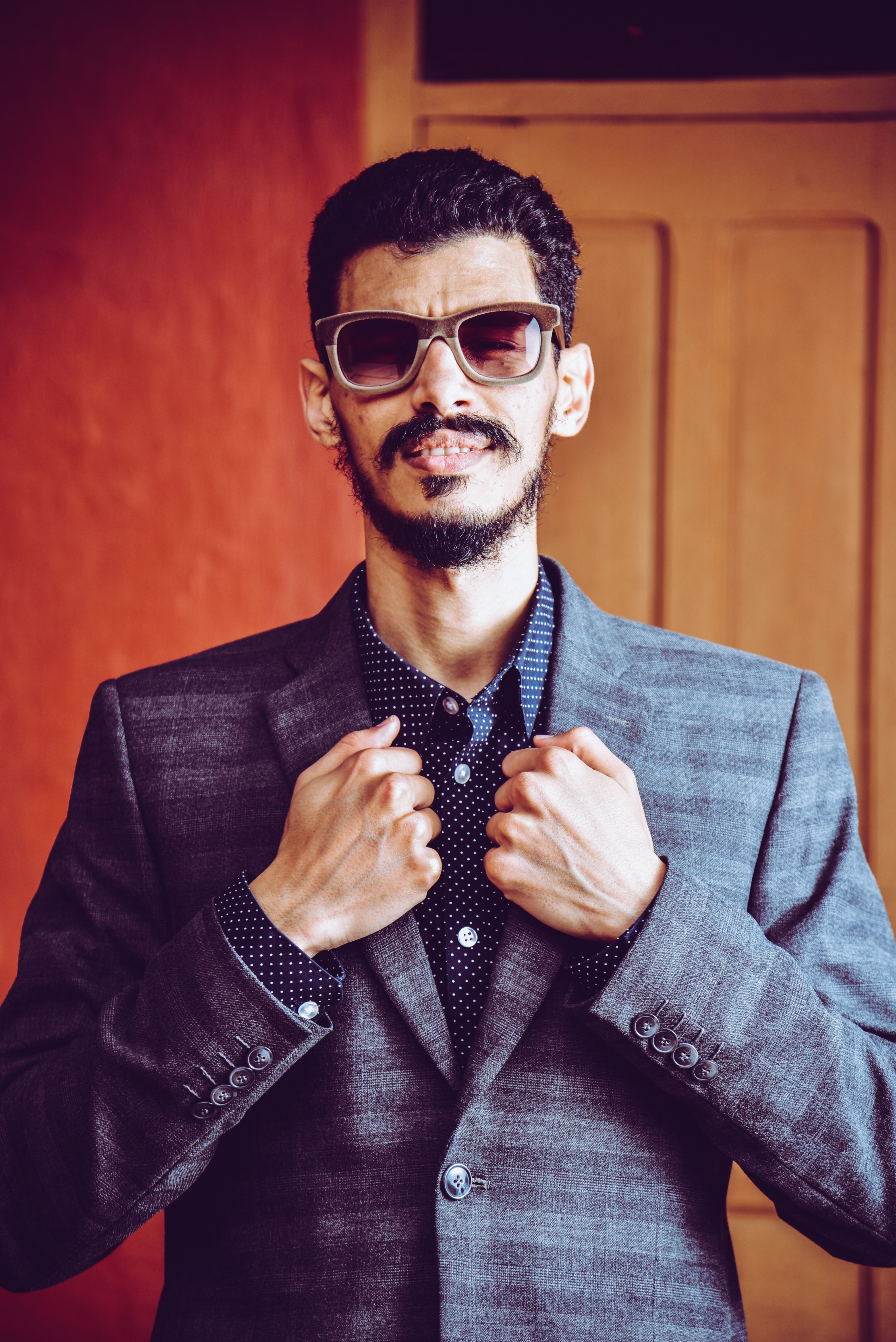 Confident man in a formal suit and sunglasses posing indoors against a warm background.