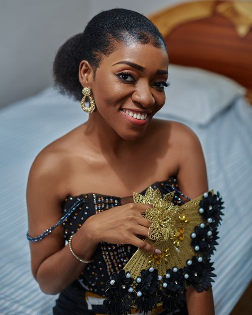 Free Smiling African woman in an off-shoulder dress holding a decorative fan indoors. Stock Photo