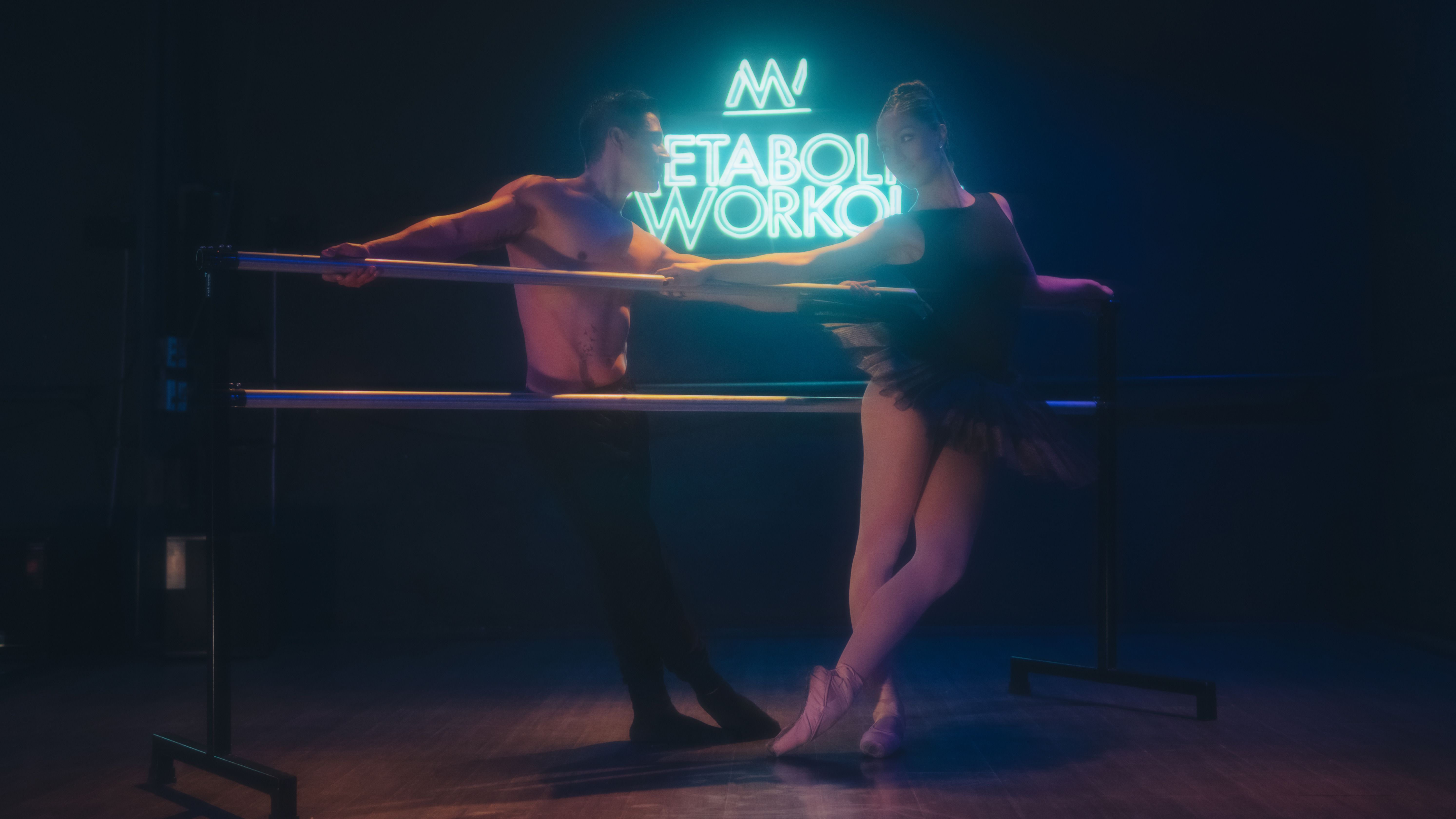 A male and female ballet dancer practicing at the studio bar under neon lights.
