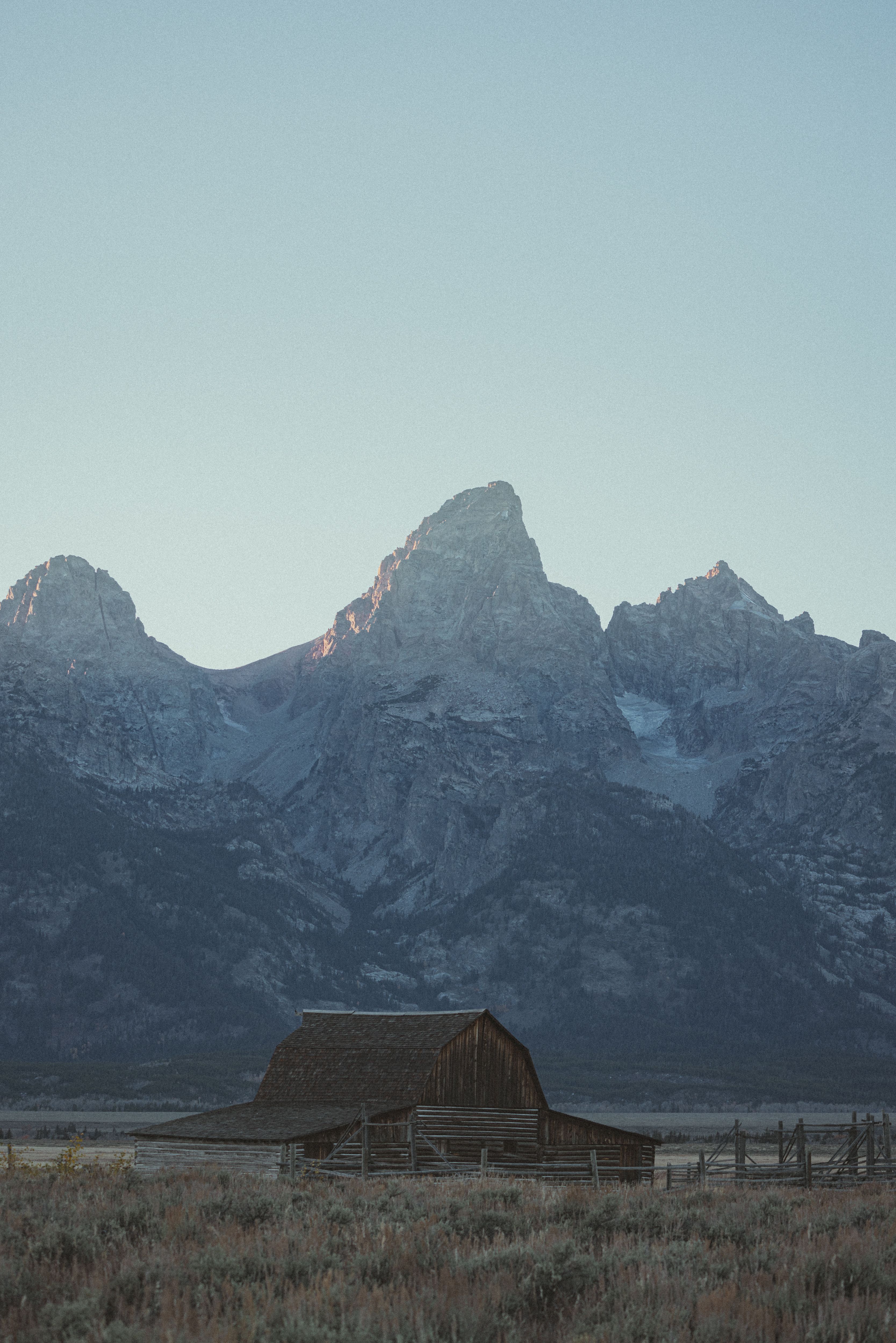 A picturesque view of a rustic barn set against stunning mountain peaks, under a clear sky.