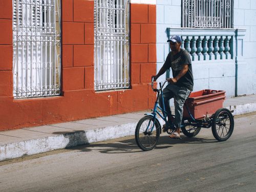Free A man rides a tricycle cart along a vibrant city street with colorful building exteriors. Stock Photo