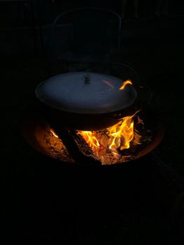 Close-up of a cooking pot over a campfire, highlighting vibrant flames at night.