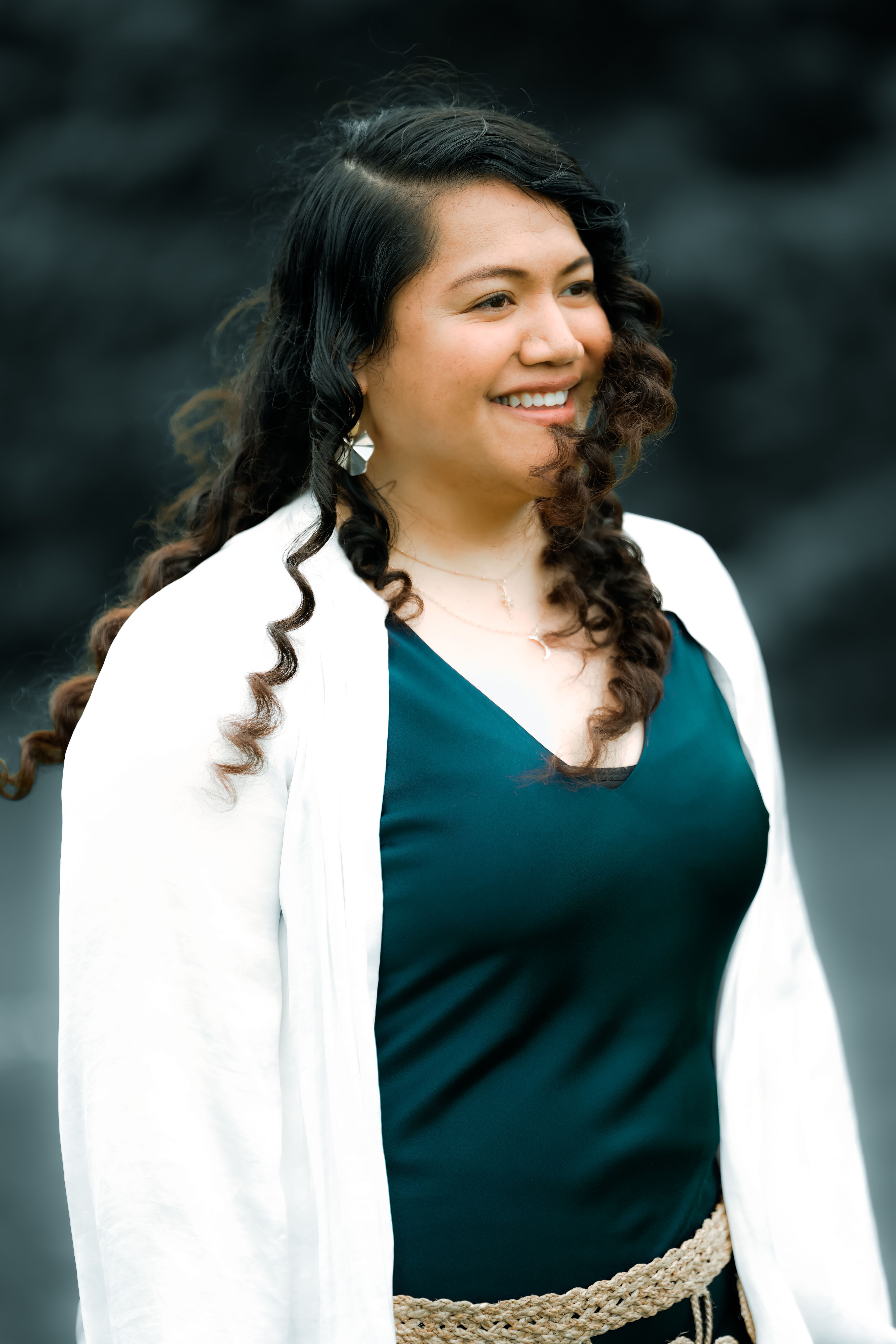 Portrait of a smiling woman with curly hair in a white blazer and blue top outside.