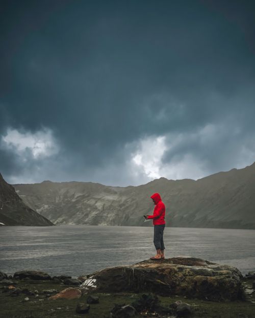 Free A person in a red jacket stands alone by a serene mountain lake under a dramatic cloudy sky. Stock Photo