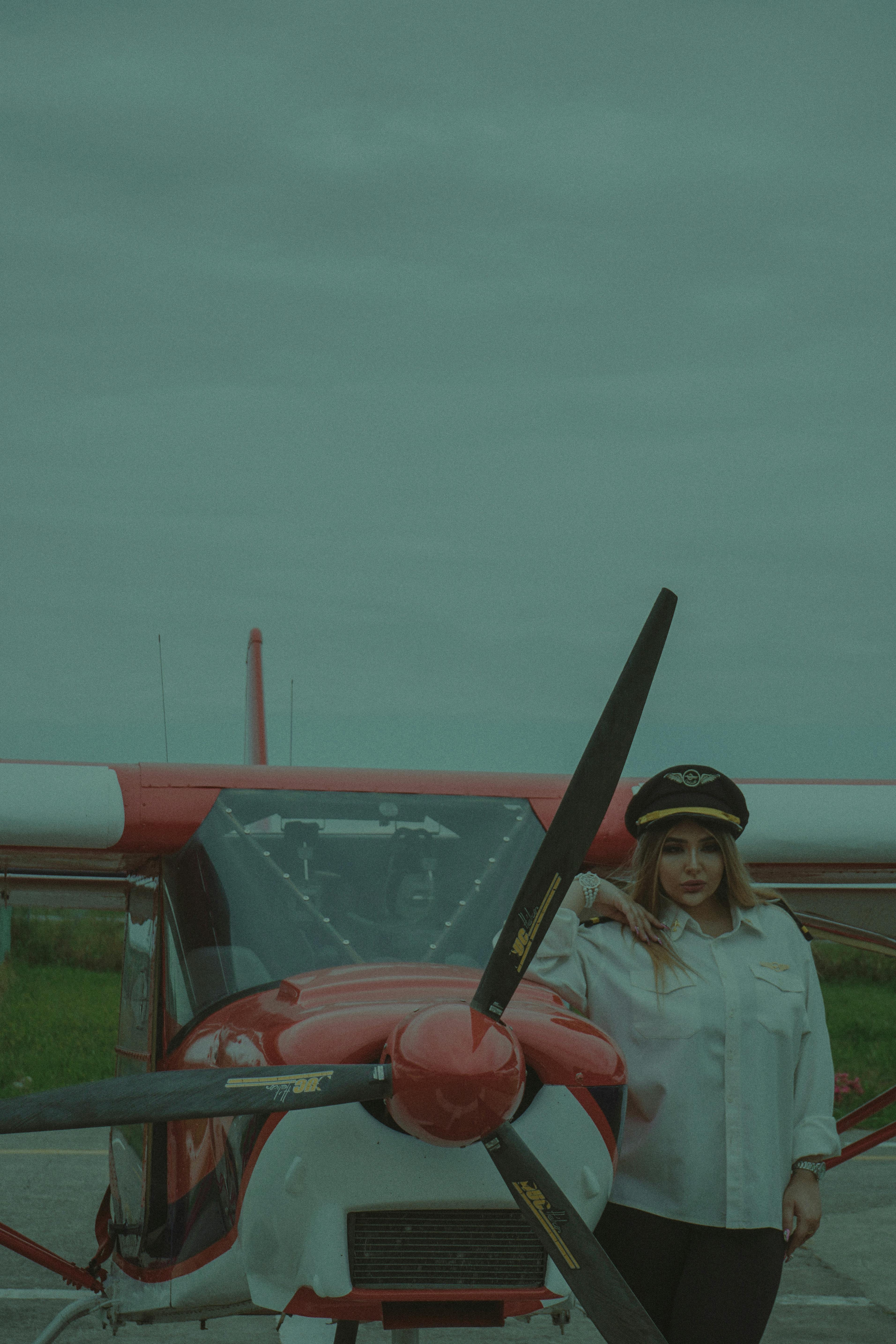 Fashionable woman pilot in uniform standing confidently by a small red aircraft on a cloudy day.