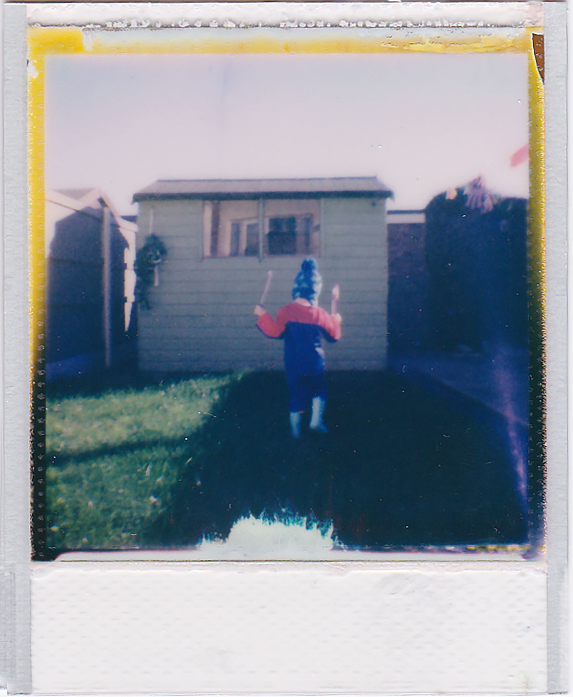 Child dressed in costume playing outside by a shed in a garden, captured on instant film.
