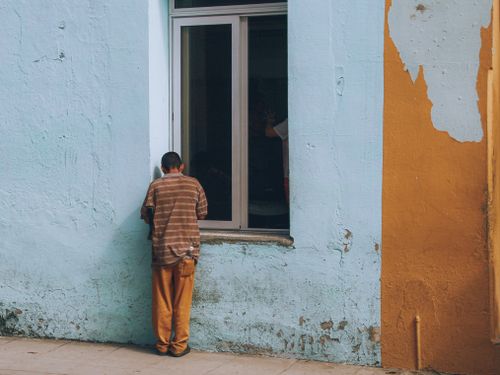 Free A man leans against a vibrant blue and orange wall, gazing into a window. Stock Photo