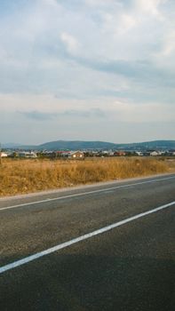 Rural landscape featuring a paved road, dry grass fields, and a cloudy sky.