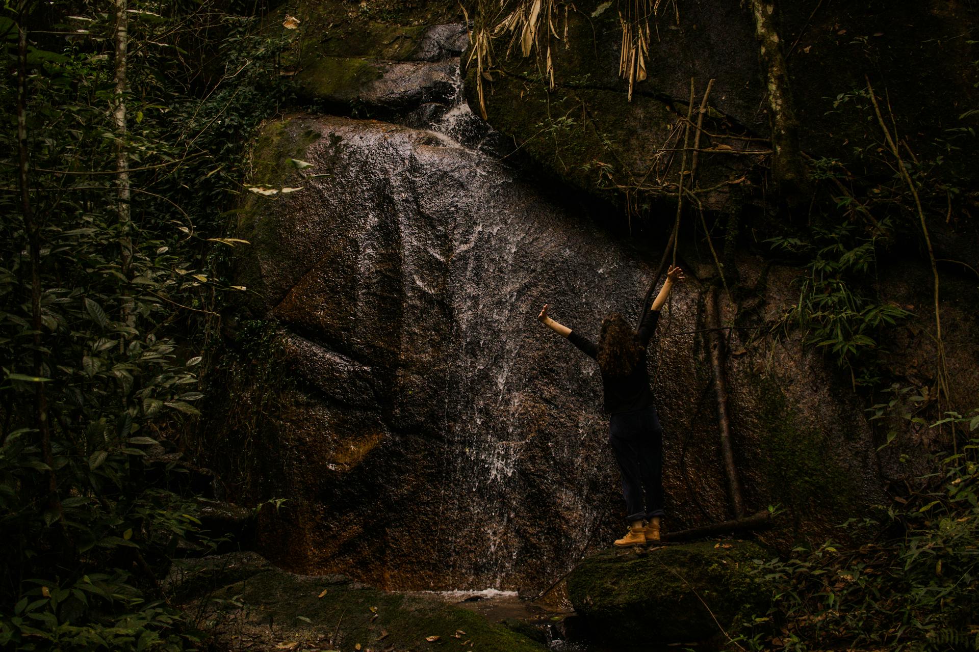 A woman raises her arms in celebration in front of a flowing waterfall in Moeda, Brazil's lush forest.