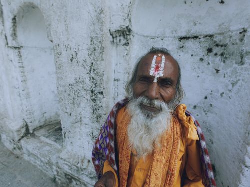 Free Smiling elderly man with a painted forehead in traditional attire against a textured wall. Stock Photo