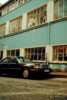 Photograph of a vintage black car parked in front of a blue industrial building exterior.