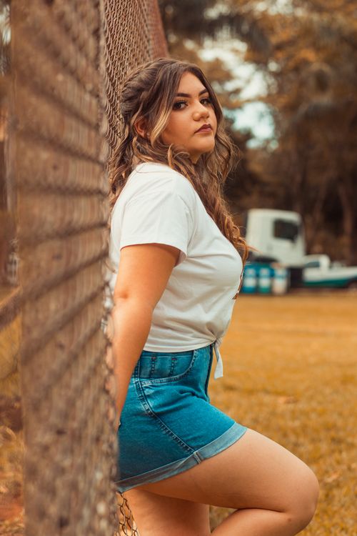 Free Young woman in white top and denim skirt leaning against a metal fence outdoors. Stock Photo