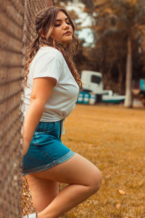 Free Stylish young woman posing near a chain link fence in a casual outfit, surrounded by greenery. Stock Photo