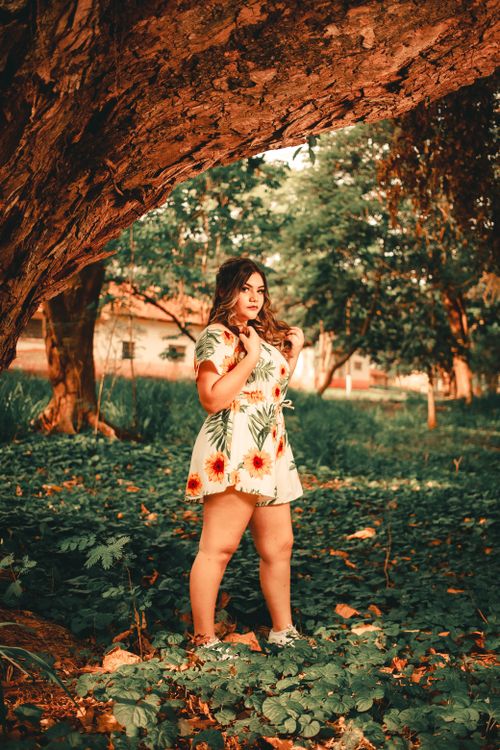 Free Fashionable woman poses under tree in a vibrant floral romper surrounded by lush greenery. Stock Photo