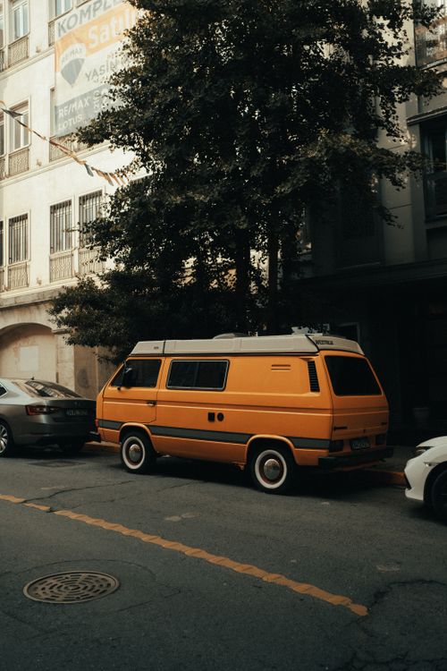 Free Orange van parked on a city street surrounded by buildings and trees. Urban transportation scene. Stock Photo